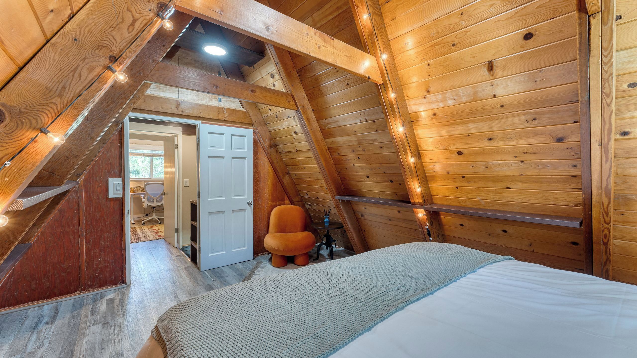 A loft bedroom with a sloped ceiling, wooden walls, a bed, a chair, and a desk with a chair.