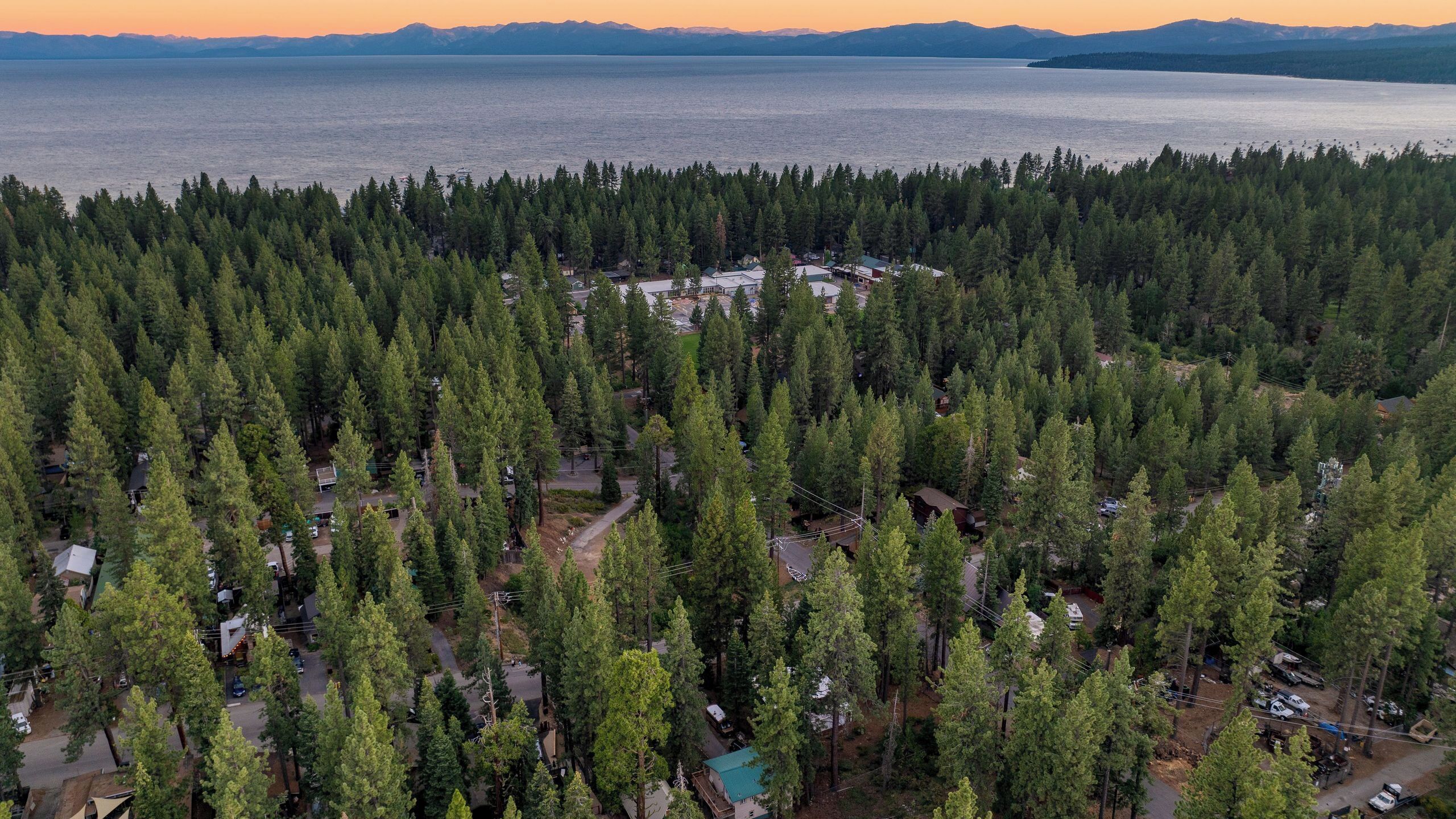 An aerial view of a forested area with a small cluster of buildings, likely a resort or lodge, surrounded by dense trees and a large body of water in the background.