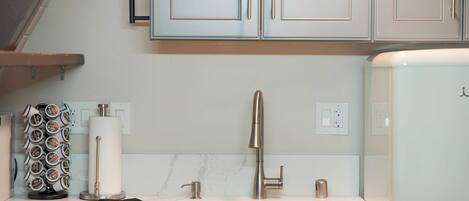 A kitchen with light-colored cabinets, a stainless steel faucet, a paper towel dispenser, and a coffee machine.