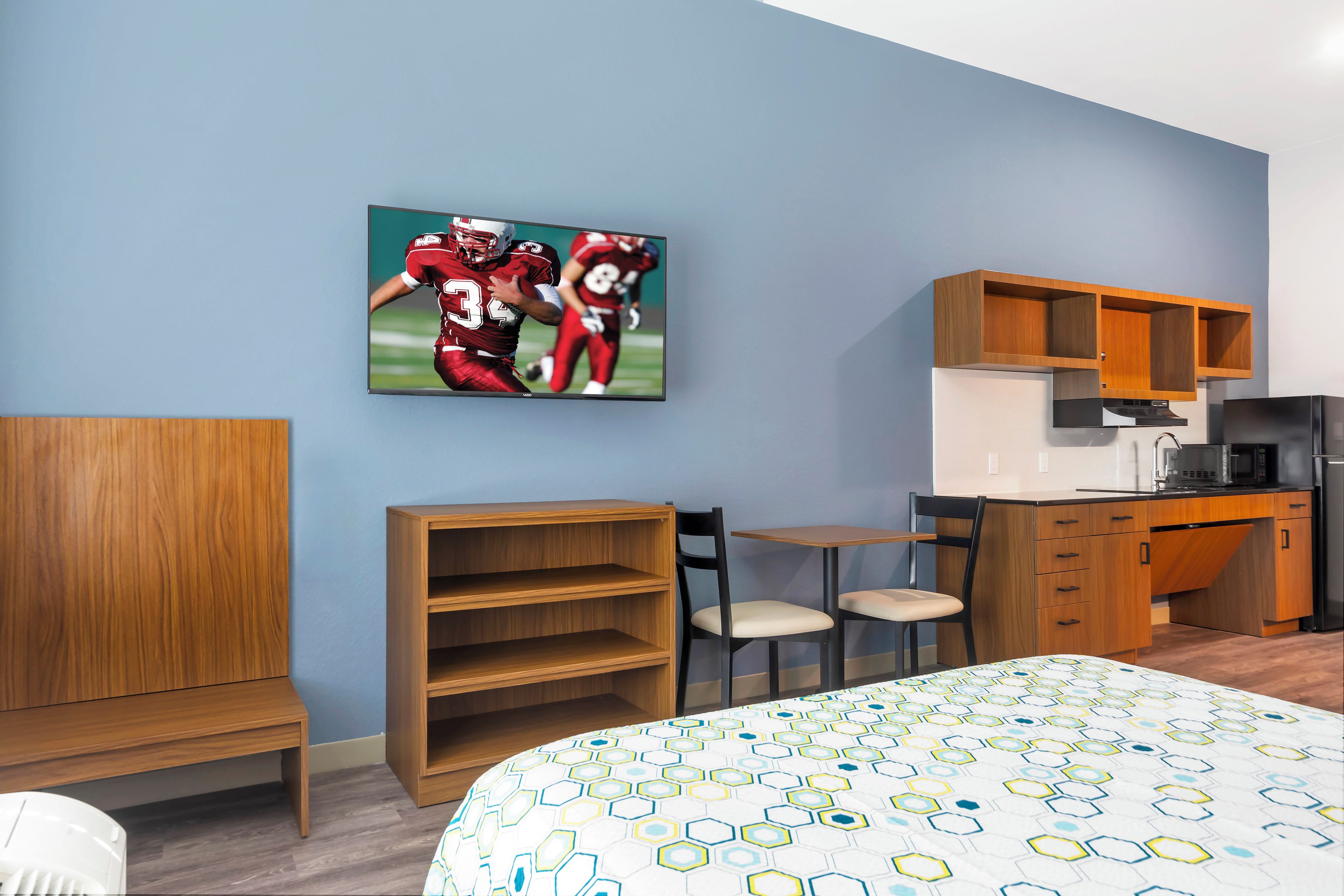 A hotel room with a bed, a desk, a chair, a TV mounted on the wall, and a kitchen area.