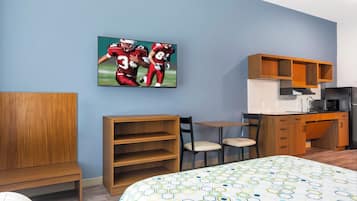 A hotel room with a bed, a desk, a chair, a TV mounted on the wall, and a kitchen area.