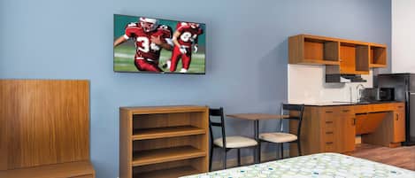 A hotel room with a bed, a desk, a chair, a TV mounted on the wall, and a kitchen area.