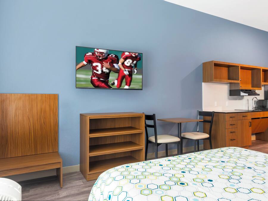 A hotel room with a bed, a desk, a chair, a TV mounted on the wall, and a kitchen area.