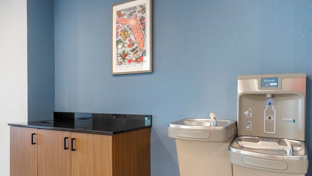 A kitchenette with a black countertop and wooden cabinets, a water dispenser, and a framed map on the wall. - HomeTowne Studios by Red Roof Tampa - Airport (Tampa)