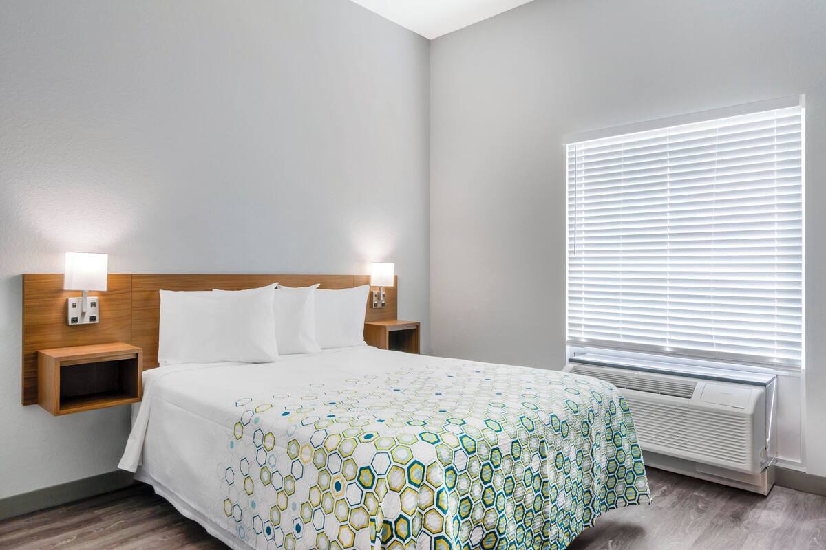 Habitación de hotel con cama, mesitas de noche, lámparas fijadas a la pared, ventana con persianas y aire acondicionado instalado en la pared.