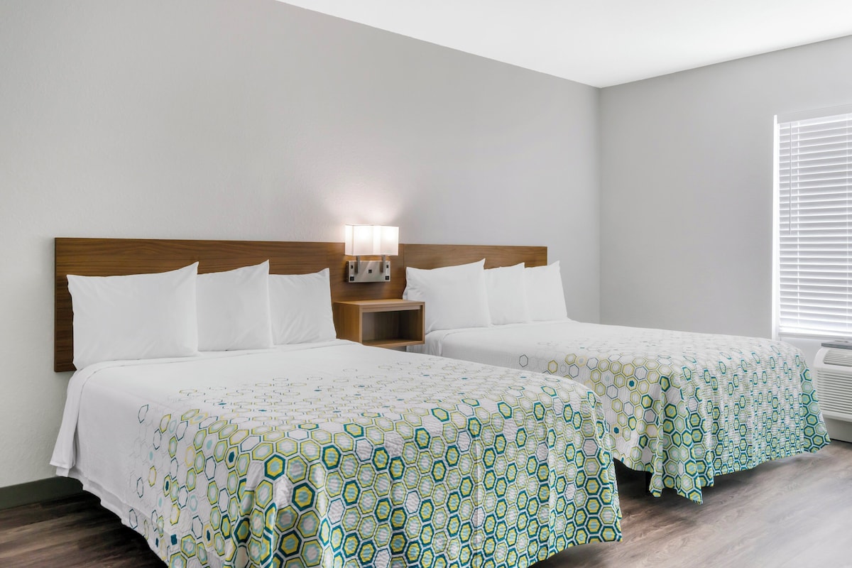 Una habitación de hotel con dos camas, una mesita de noche con lámpara y una ventana con persianas.