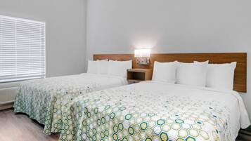A hotel room with two beds, a wooden headboard, a nightstand with a lamp, and a window with blinds.