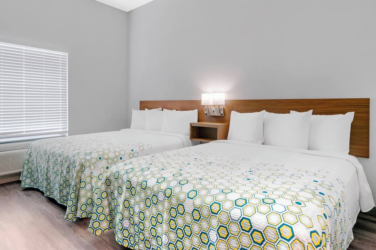 Habitación de hotel con dos camas, cabecera de madera, mesita de noche con lámpara y ventana con persianas.