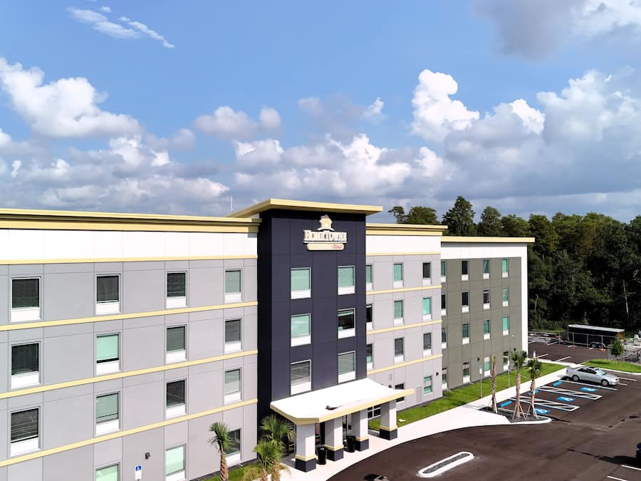 A multi-story hotel with a central entrance, parking lot, and a clear sky with clouds.
