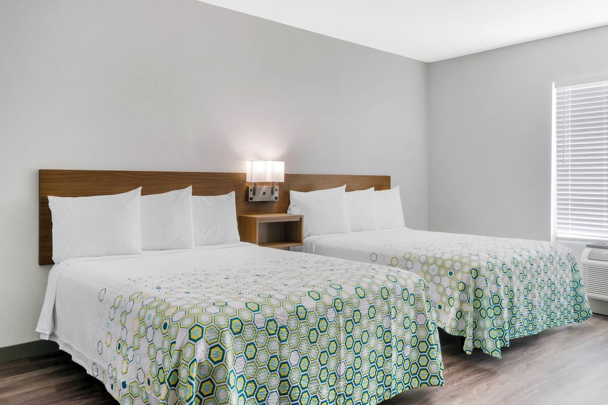 Una habitación de hotel con dos camas, una mesita de noche con lámpara y una ventana con persianas.