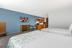Habitación de hotel con dos camas, un escritorio, una silla, una estantería y una zona de cocina.