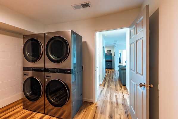 A laundry room with stacked washers and dryers, a wooden floor, and a hallway leading to a kitchen area.