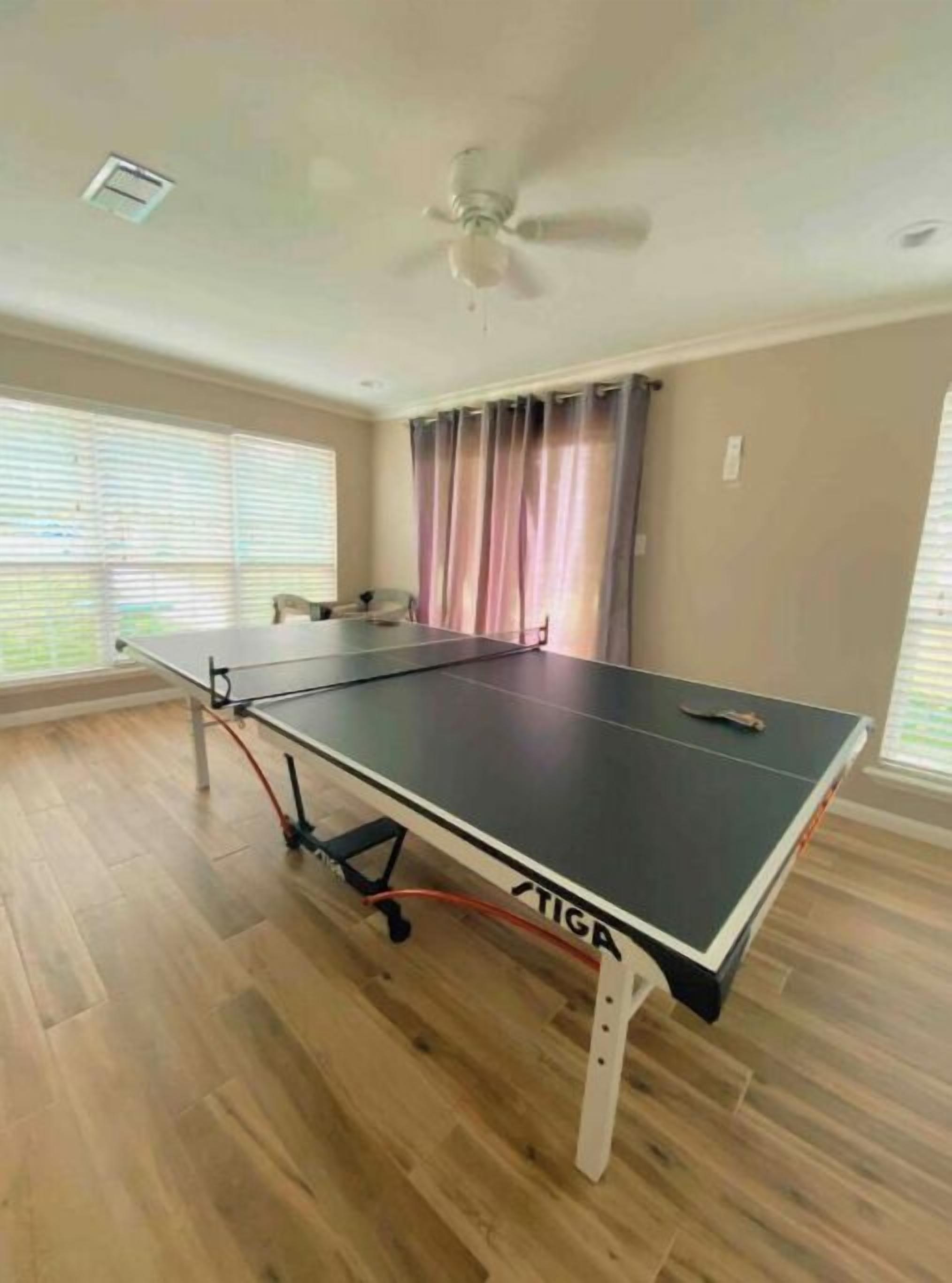 A room with a ping pong table, a ceiling fan, and large windows with blinds.
