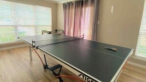 A room with a ping pong table, a ceiling fan, and large windows with blinds.