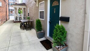 A narrow alleyway with a wooden table and chairs, two potted plants, a green door with the number 83, and a 'hello' mat. -  The Lounge - Downtown Chillicothe (Chillicothe)