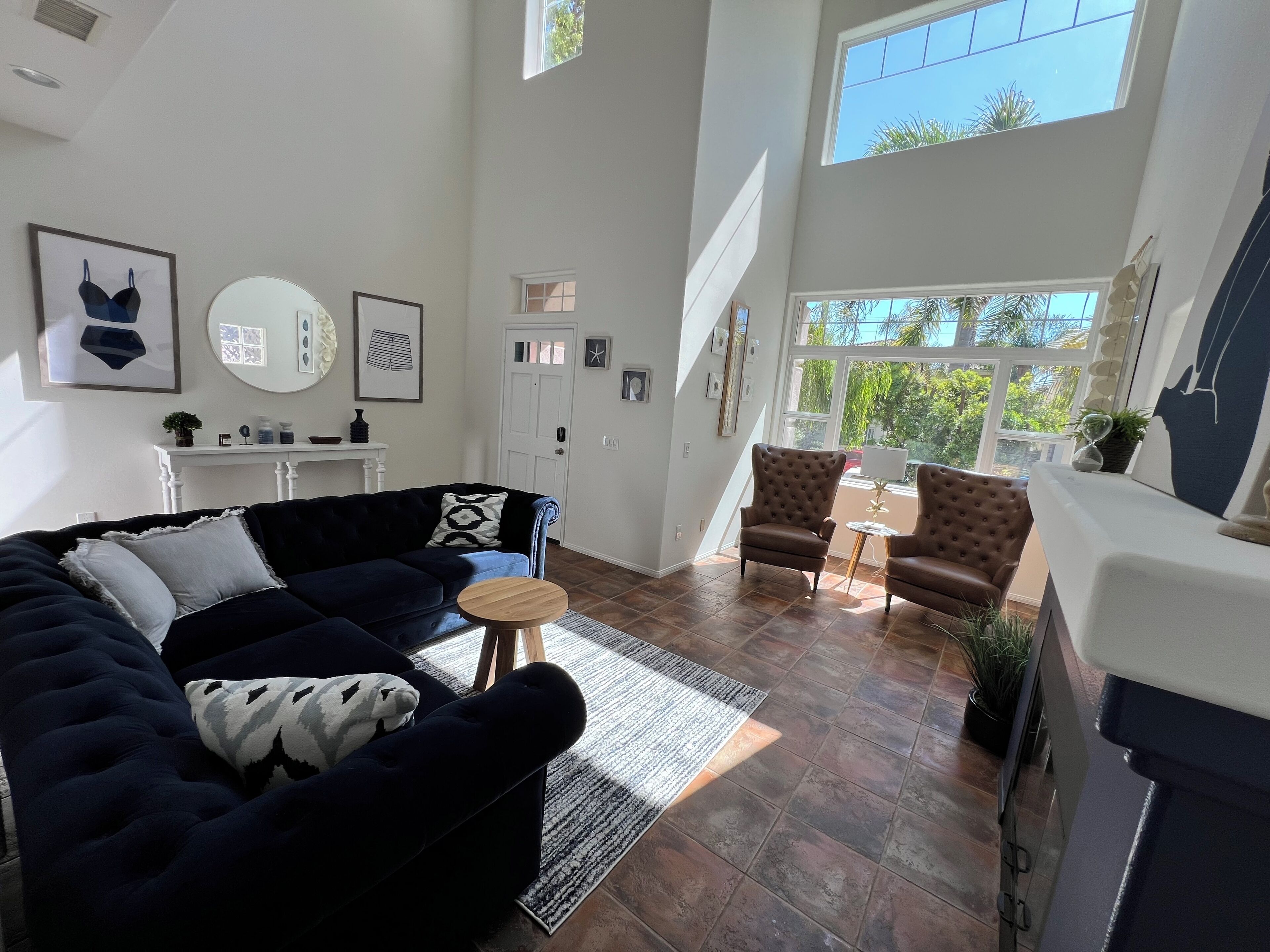 A spacious living room with a dark blue sofa, two brown armchairs, and a small round table. There are large windows providing natural light and a view of greenery outside.