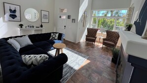 A spacious living room with a dark blue sofa, two brown armchairs, and a small round table. There are large windows providing natural light and a view of greenery outside. - OCEANSIDE BEACH HOUSE~Dreamy Palm Tree Lined Street~5 MIN WALK to EVERYTHING!  (Oceanside)