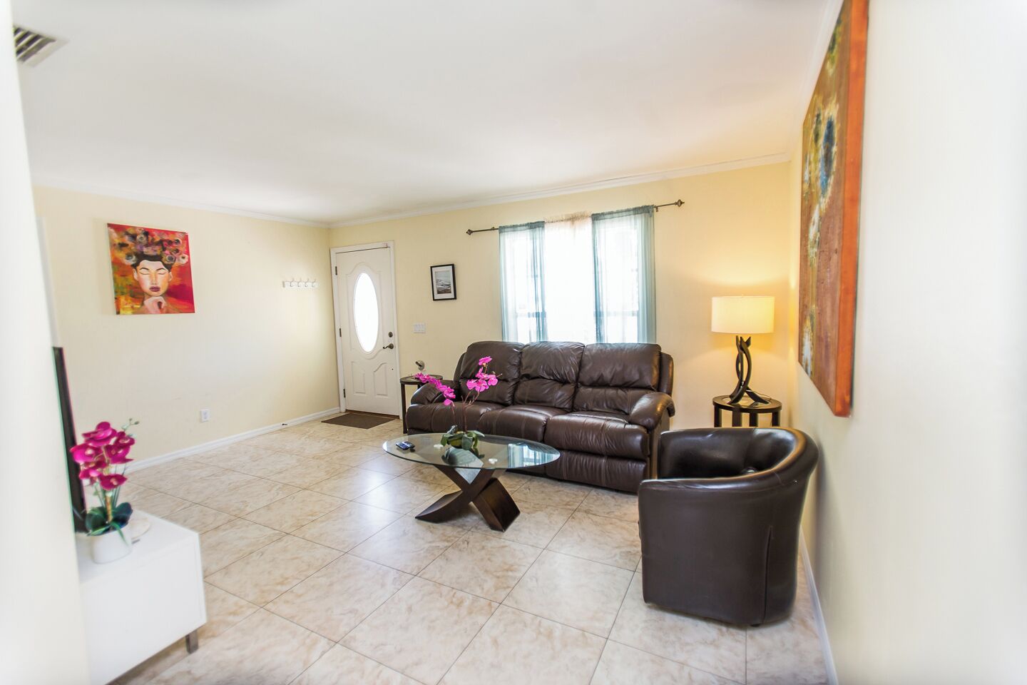 A living room with a brown leather sofa, a glass coffee table, a white side table, and a black armchair. There is a door, a window with curtains, and a painting on the wall.