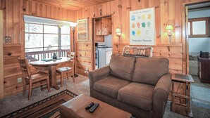 A cozy cabin interior with wooden walls, a sofa, a dining table with chairs, a kitchen area, and a large window. - Get Cozy on Rainbow, 1 Night Free! (Big Bear City)