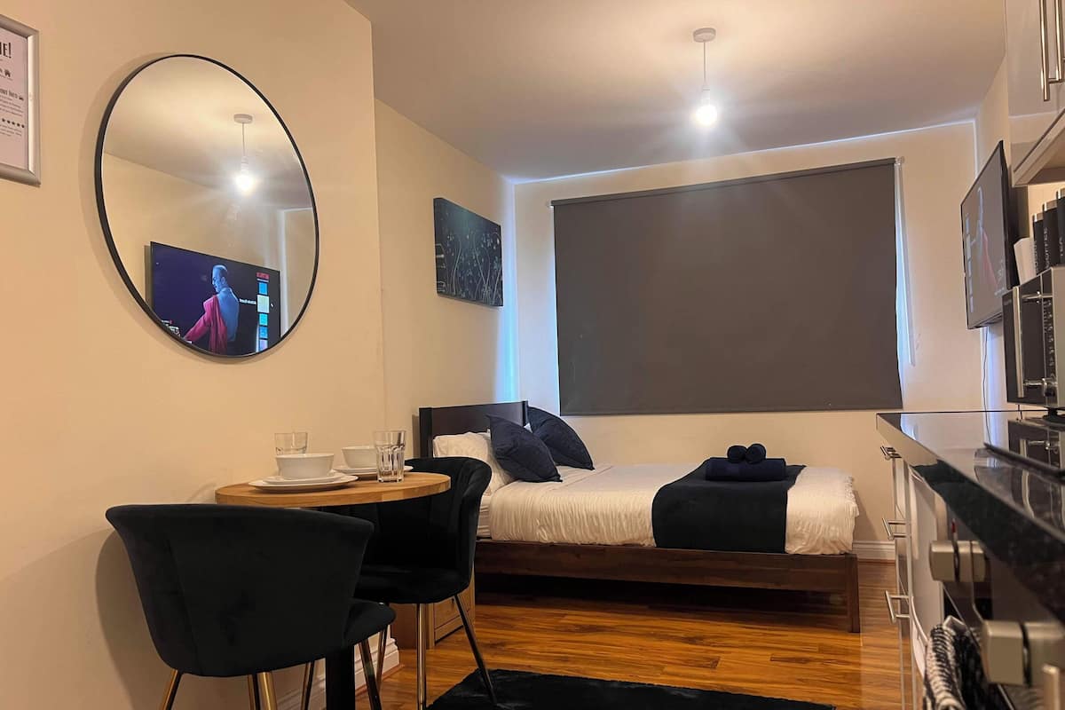 A compact hotel room with a bed, a round mirror, a small table with chairs, a television, and a kitchenette.