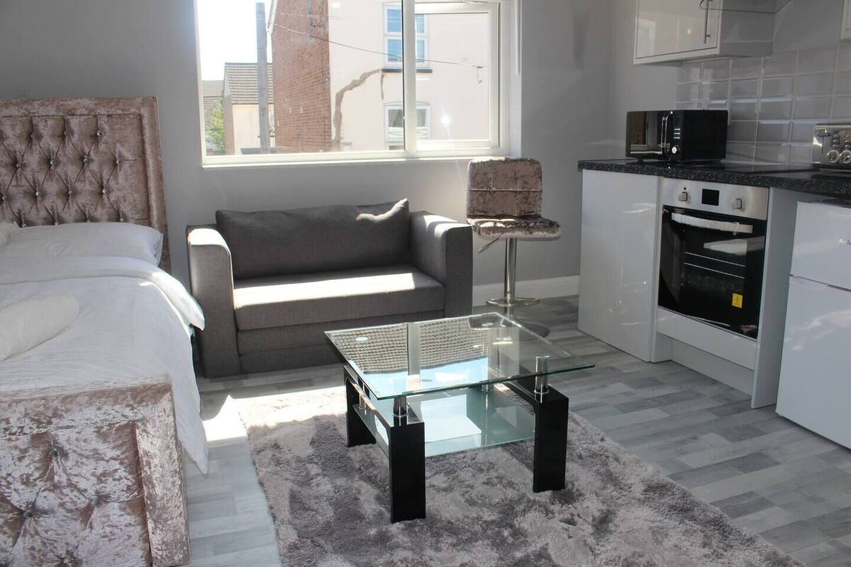A modern bedroom with a bed, a grey sofa, a glass coffee table, and a kitchen area with a microwave and oven.