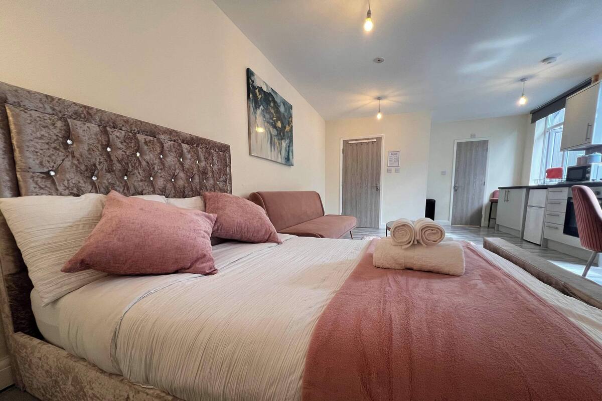 A neatly arranged bedroom with a large bed, a headboard with decorative lights, a sitting area with a chair, and a view of the kitchen through an open door.