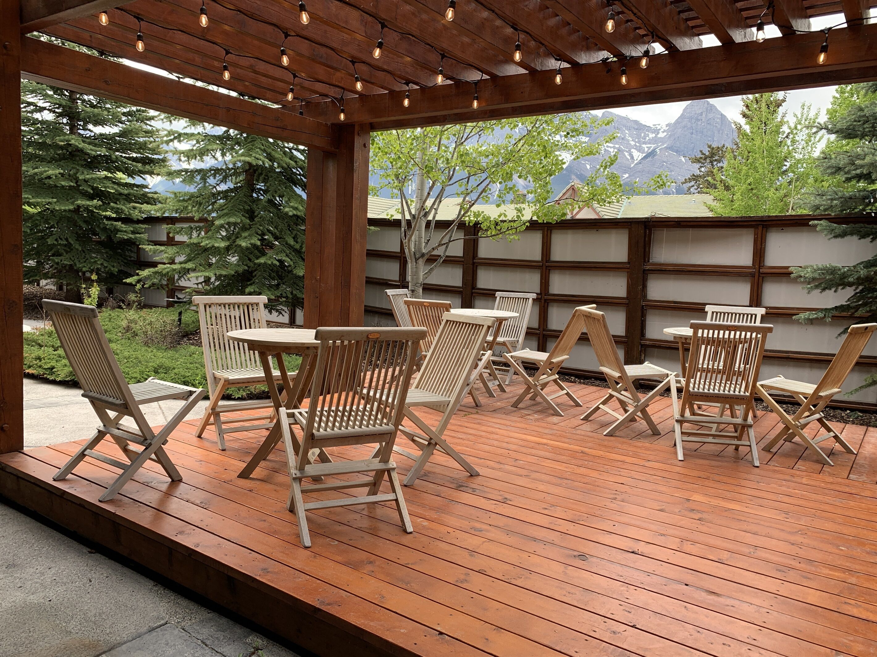 An outdoor patio with wooden chairs and tables, a wooden pergola with string lights, and a view of mountains and trees.