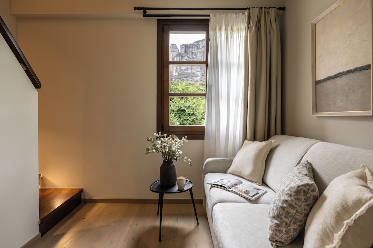 En hyggelig stue med en beige sofa, et lille sofabord med en vase og en kop, et vindue med gardiner, og et billede i ramme på væggen.
