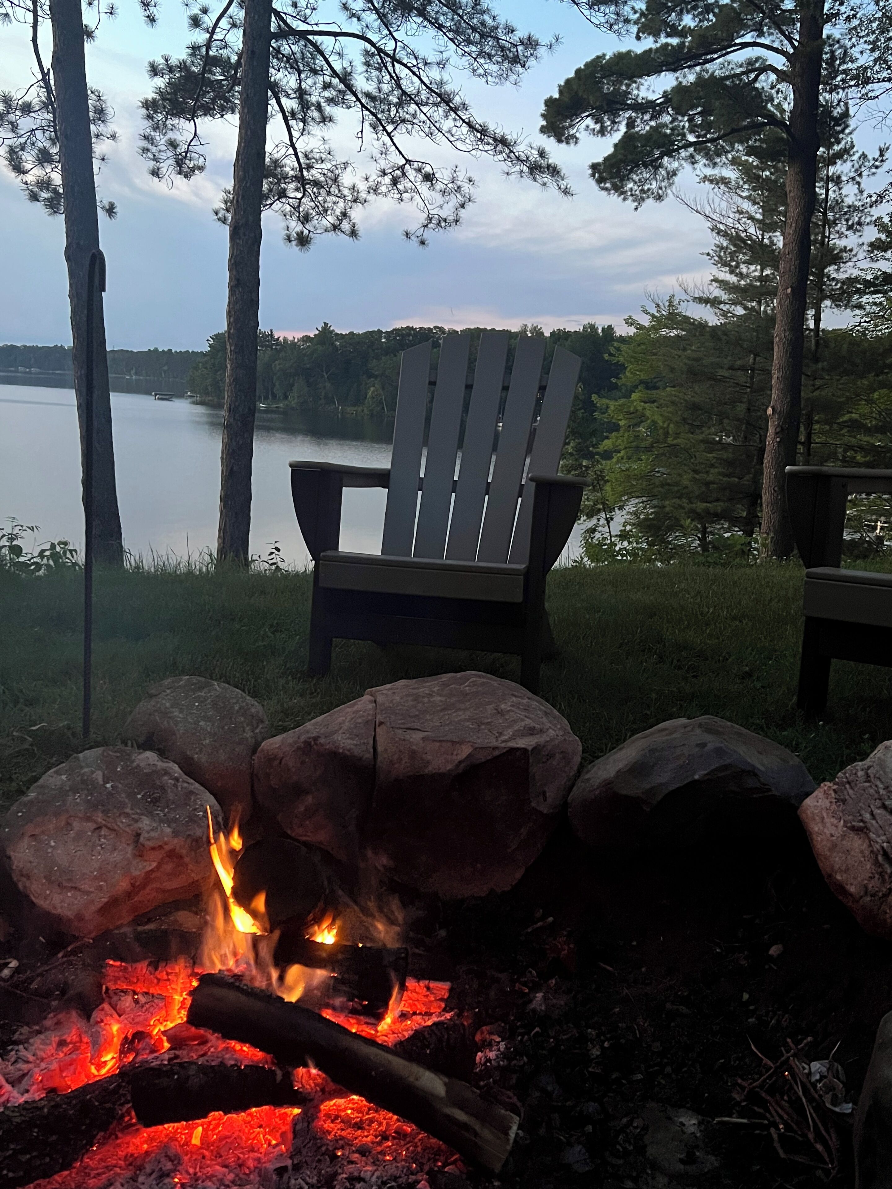 A campfire with rocks, a wooden chair, and a view of a lake surrounded by trees.