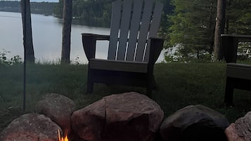 A campfire with rocks, a wooden chair, and a view of a lake surrounded by trees.