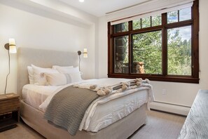 A bedroom with a large bed, two bedside lamps, a window with a view of trees, and a wooden nightstand.