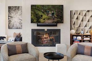 A living room with a fireplace, a television mounted above it, and a bookshelf with various items. - Charlie's Slopeside: Bachelor Gulch Ski-In/Out, Sleeps 8, Ritz Pool Access + Slopeside Hot Tubs (Avon)