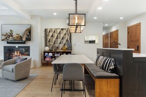 A modern kitchen with a central island, a dining area with a long table and chairs, a sofa with patterned cushions, and a fireplace.