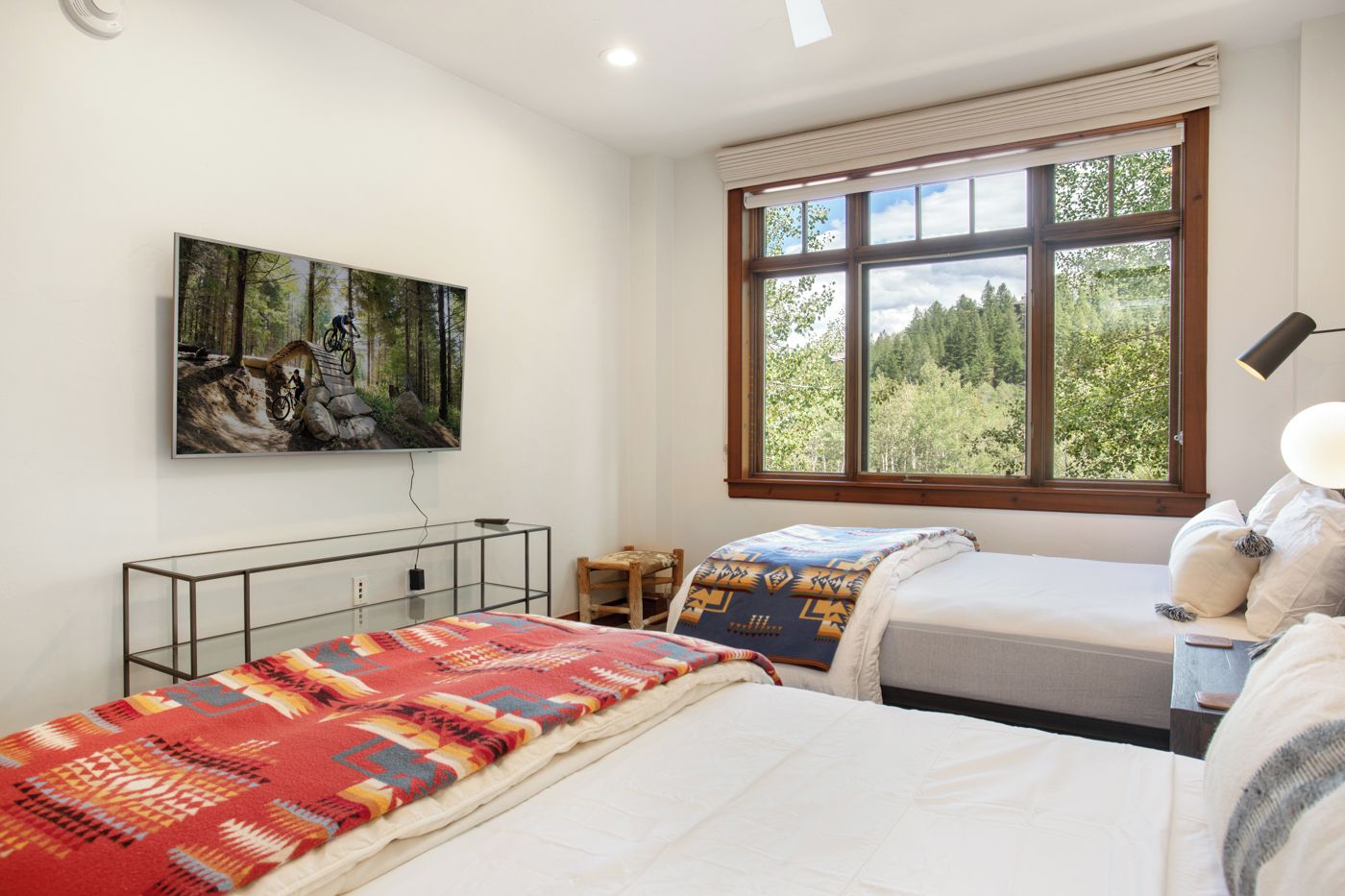 A bedroom with two beds, a large window with a forest view, a mounted painting, and a bedside table with a lamp.