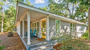 Une maison de plain-pied dotée d’un porche couvert, d’une porte d’entrée et d’un petit jardin devant, avec un banc et des chaises.