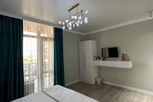 Un dormitorio con cama, armario, televisor en una estantería y balcón con cortinas.