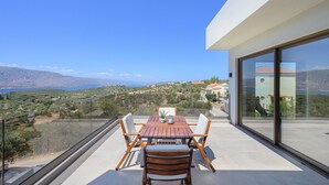 Un balcon avec une table et des chaises en bois, donnant sur un paysage pittoresque composé de collines et d’une étendue d’eau.