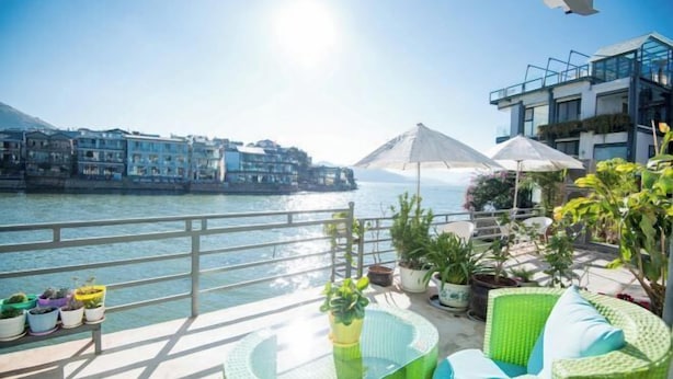 A balcony with a glass table, green wicker furniture, potted plants, and a view of a body of water and buildings. - Lanyun Haiwan Inn (Dali City)