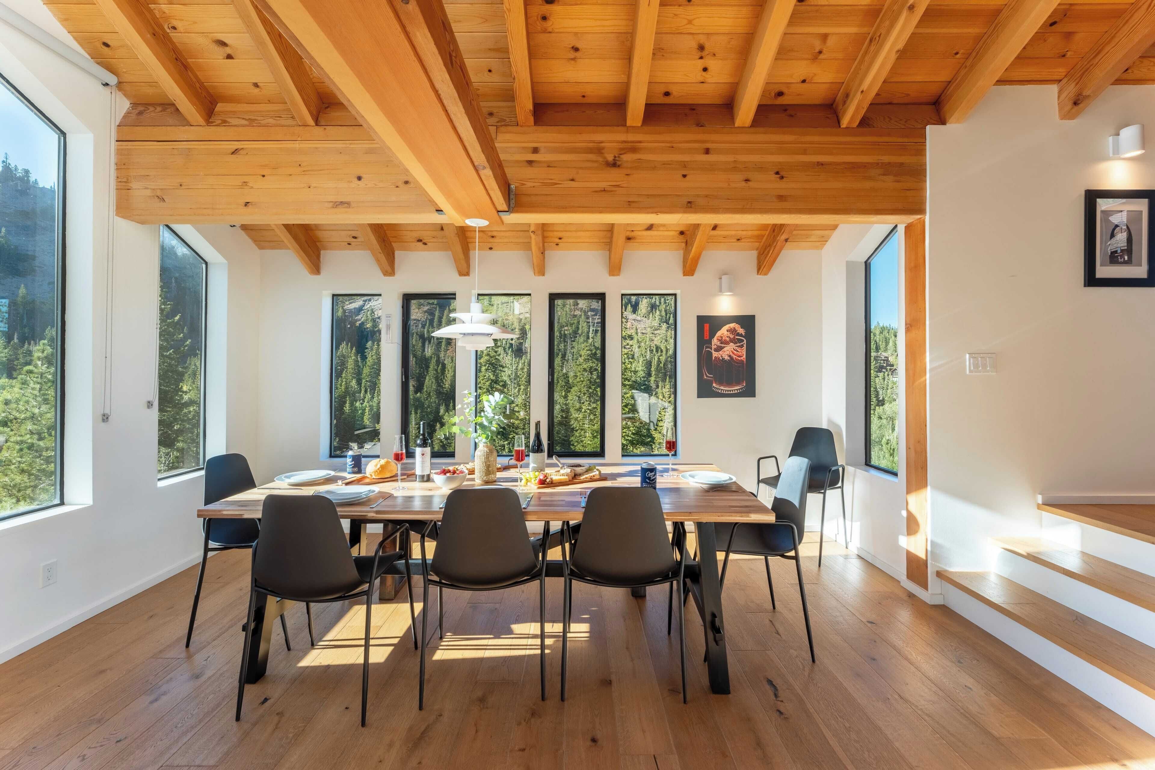 A dining area with a wooden table, chairs, and a view of trees through large windows.