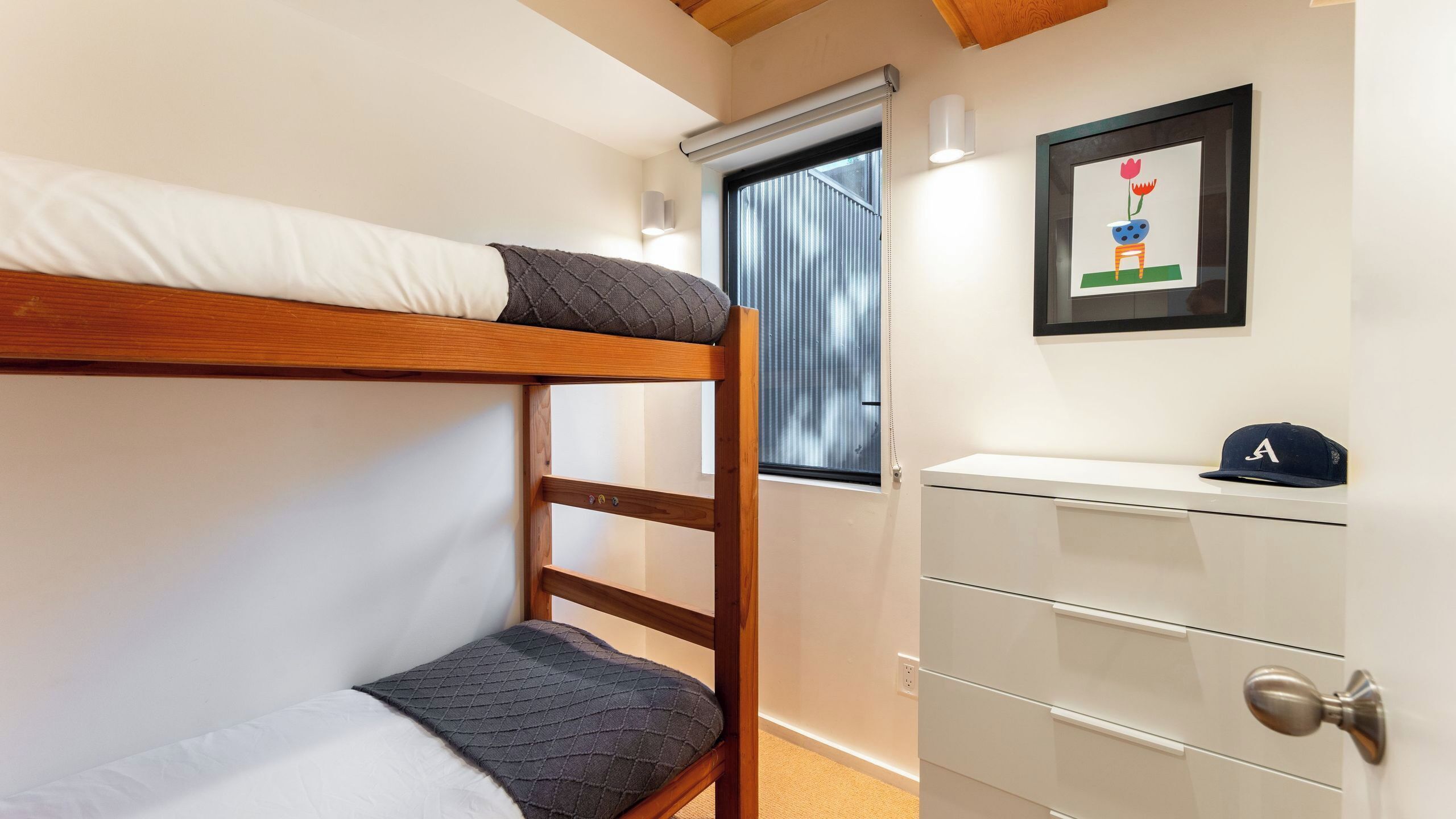 A bunk bed with a pillow, a white dresser with drawers, a framed picture on the wall, and a window with blinds.