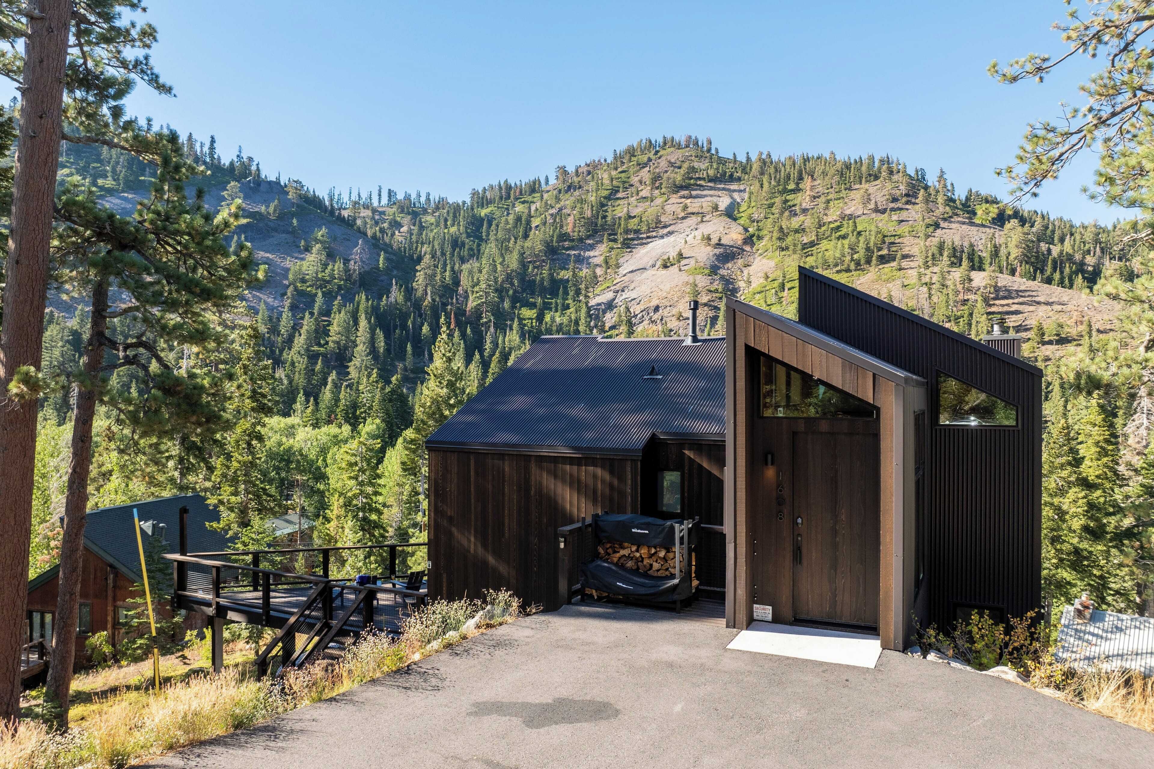 A modern cabin with a wooden exterior, a covered outdoor seating area, and a view of a forested mountain.