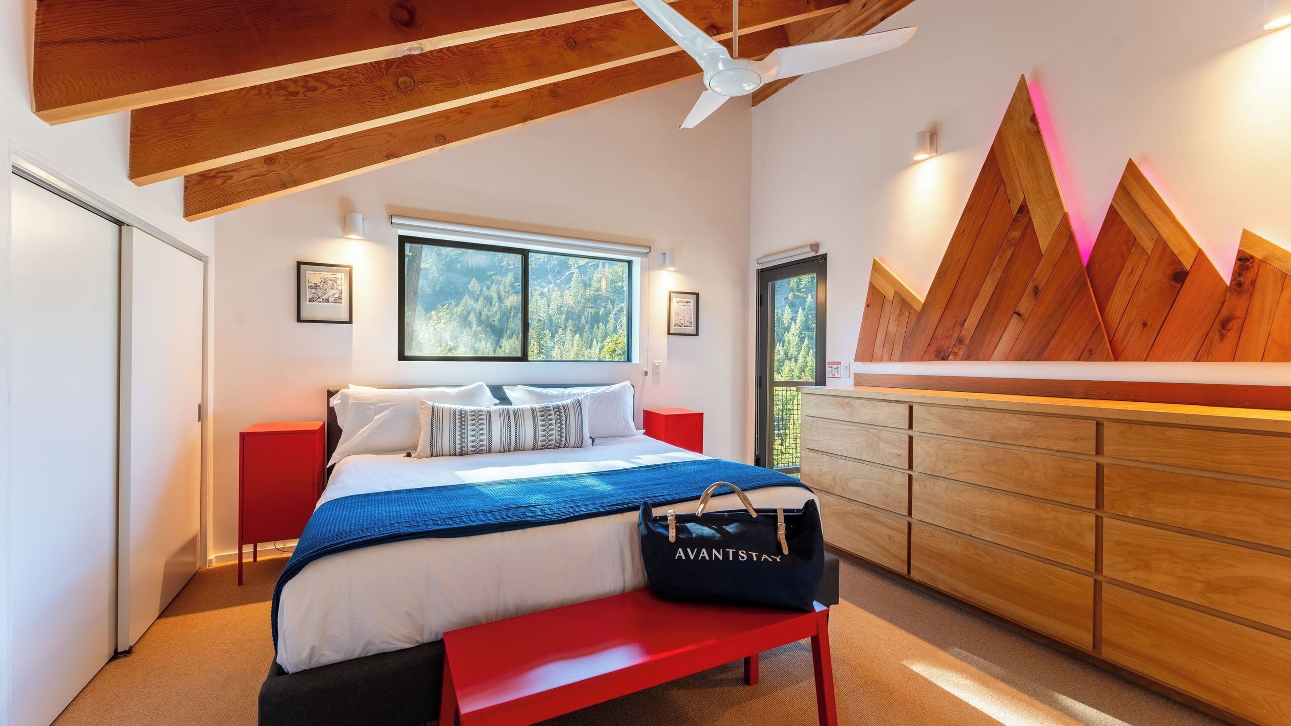 A modern bedroom with a bed, a red bench, a wooden dresser, and a large window with a view of trees.