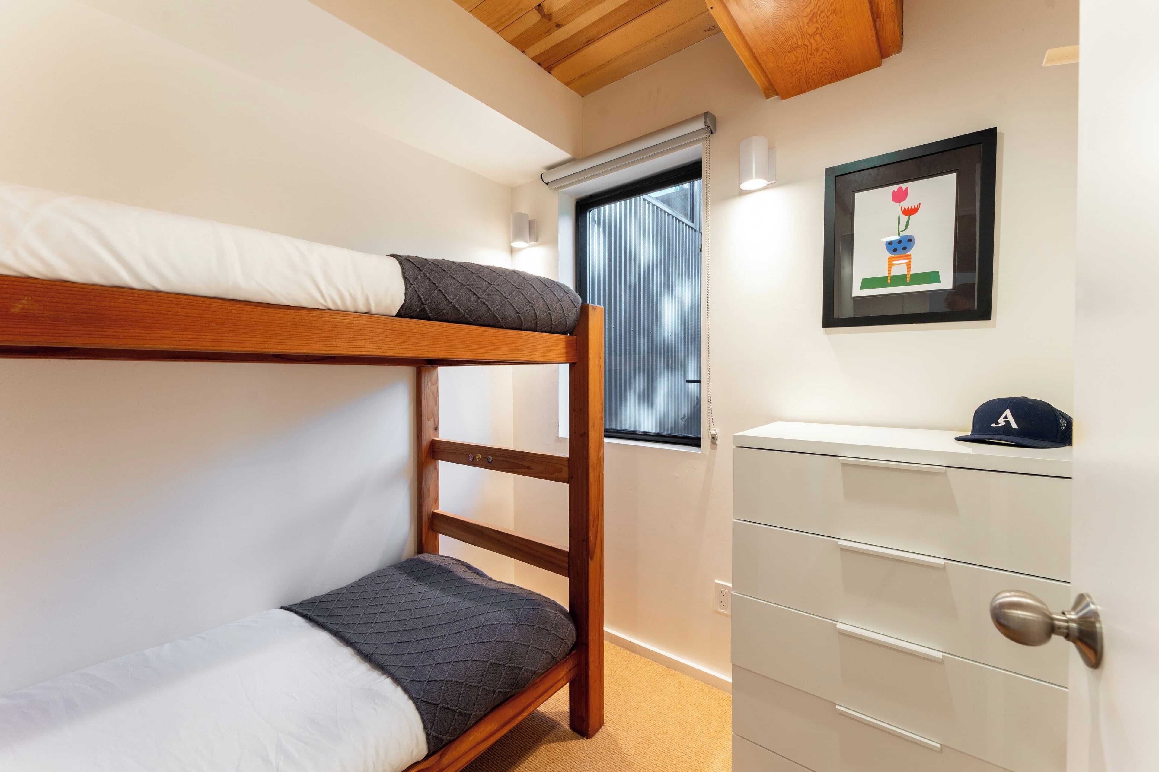 Een stapelbed met houten frame en een wit matras, een witte dressoir met lades, en een ingelijste foto aan de muur.