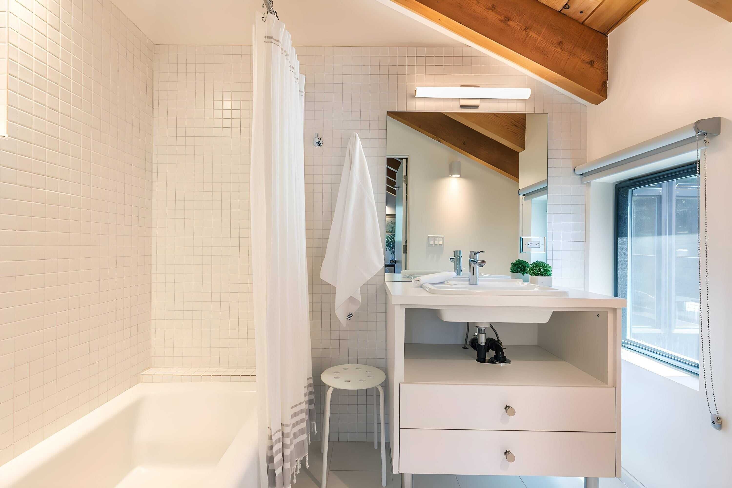 Une salle de bain moderne avec une baignoire, un meuble-lavabo blanc avec un miroir et une fenêtre laissant entrer la lumière naturelle.