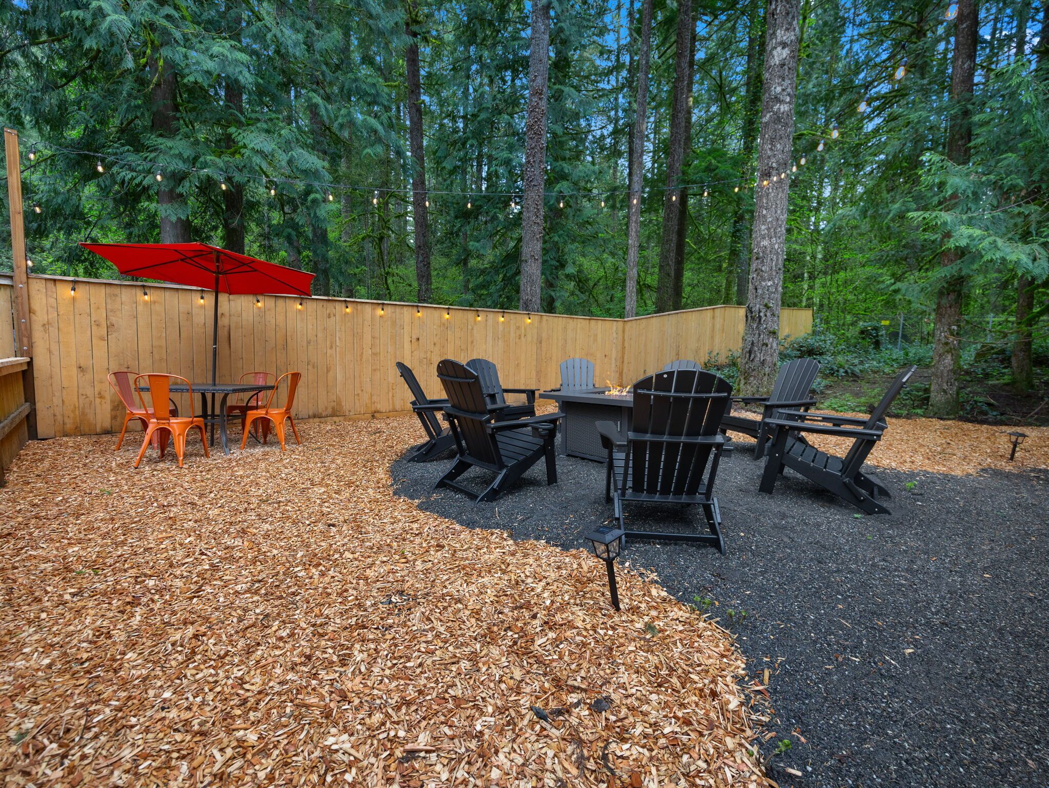 An outdoor patio with a wooden deck, Adirondack chairs, a table, and a red umbrella.
