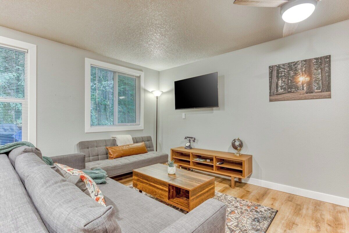 Ein Wohnzimmer mit grauem Sofa, einem Holztisch, einem an der Wand befestigten Fernseher und einem Fenster mit Jalousien.