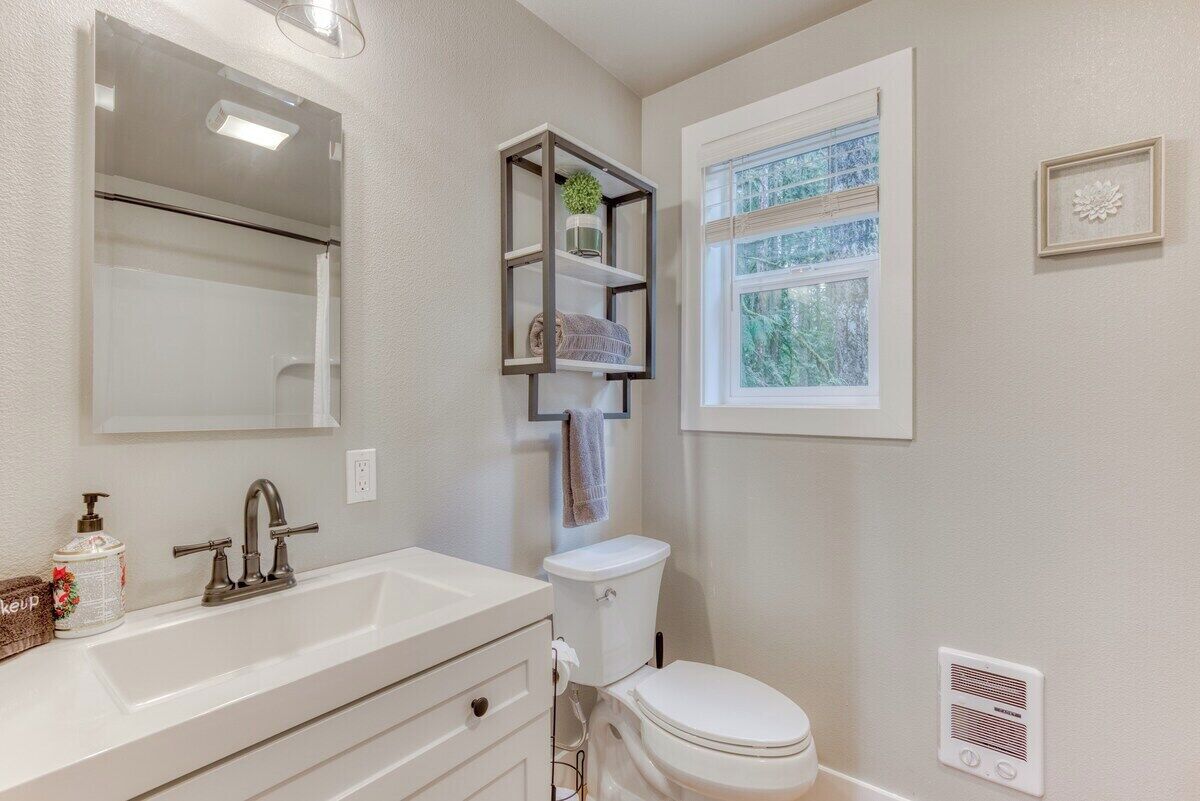 Une salle de bain avec un lavabo blanc, un miroir, des toilettes, une fenêtre et une étagère sur laquelle se trouvent une plante et une serviette.