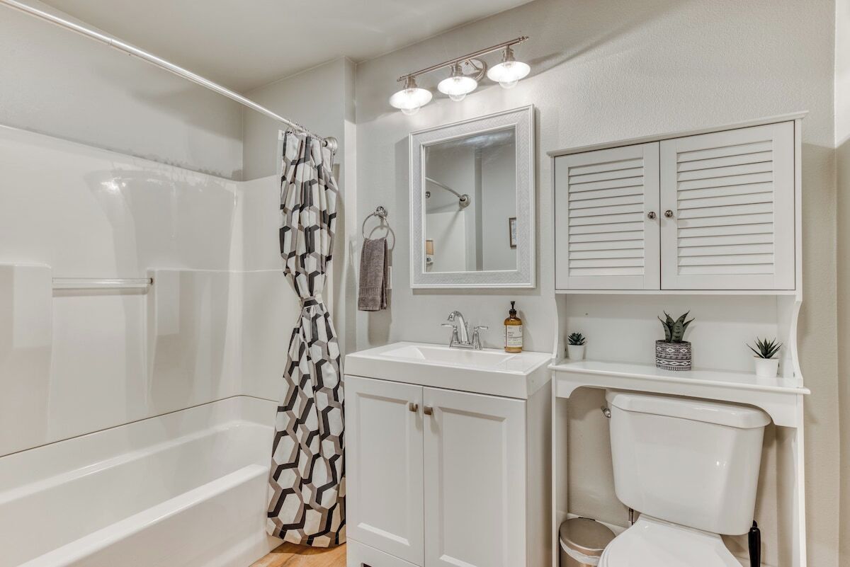 A bathroom with a bathtub, a shower curtain, a sink with a mirror above it, a toilet, and a cabinet with a shelf above the toilet.