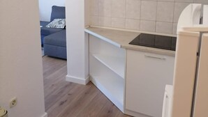 A compact kitchen with white cabinets, a countertop, and a refrigerator. A grey sofa is visible in the adjacent living area. - Apartments Libertas - One Bedroom Apartment with Terrace and Sea View - (5) (Rogoznica)