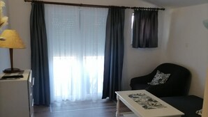 A living room with a sofa, a coffee table, and a lamp. There is a window with curtains and a view outside. - Apartments Libertas - One Bedroom Apartment with Terrace and Sea View - (5) (Rogoznica)